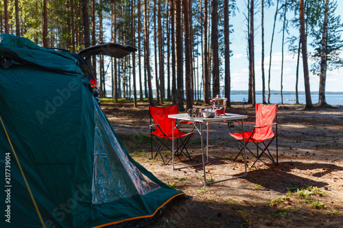 Family vacation, travel concept on wheels, a road trip, a table and chairs, a tent and a SUV with the back door open on site. Folding table and chairs with gas stove in the camping in the forest.