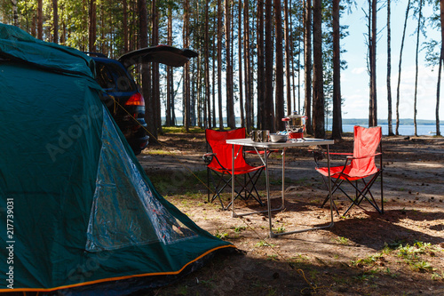 Family vacation, travel concept on wheels, a road trip, a table and chairs, a tent and a SUV with the back door open on site. Folding table and chairs with gas stove in the camping in the forest.