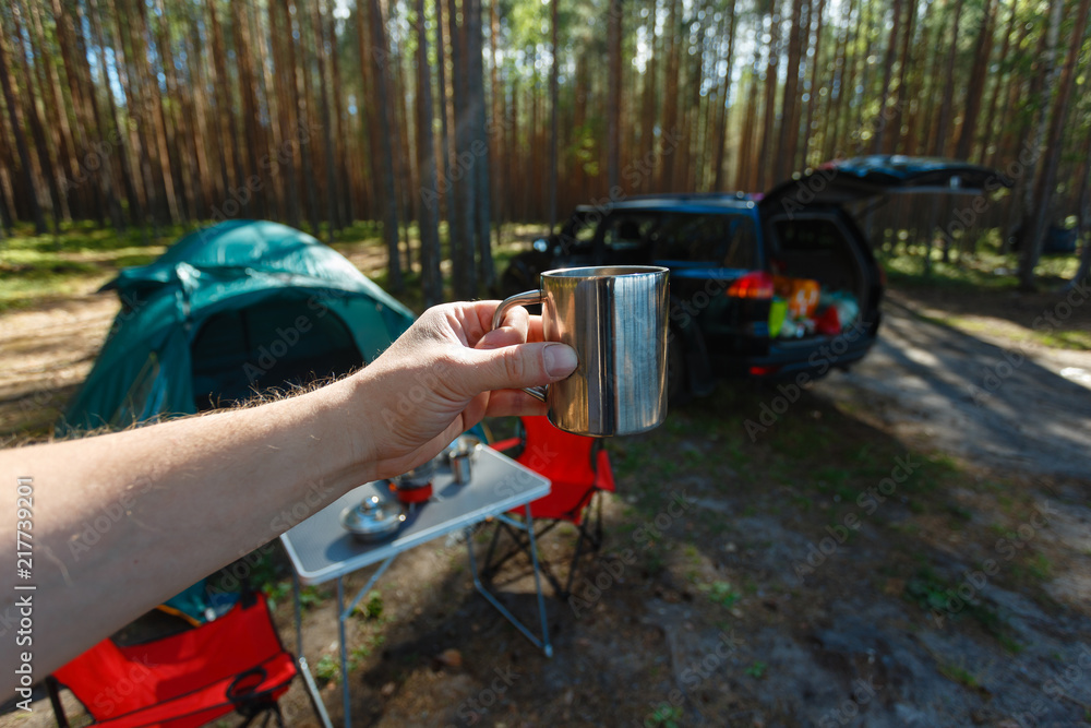 The man's hand holds an iron mug with a warm drink on the background of the table and chairs, campfire, a tent and an SUV in camping. Family vacation, concept of traveling by SUV or car in the wild.