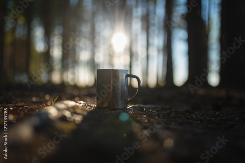 Close - up of iron camping mug with a warm drink in the hands of a male traveler on a forest and sunset background. Concept of camping in the forest. Picnic on a warm summer evening at sunset.