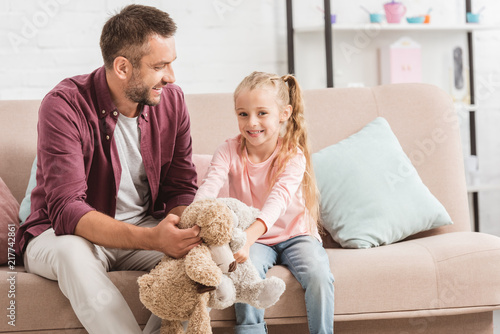 father and smiling daughter holding teddy bears, sitting on couch