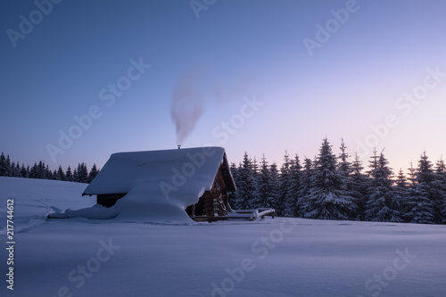 Winter landscape of a mountain forest with a cabin at sunset. Trail stamped in the snow towards the cabin. Fir-trees and a cabin covered with snow.
