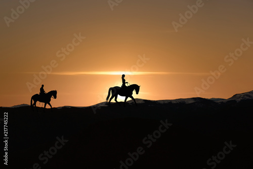 Silhouette of man and woman riding horse across horizon as the sun goes down