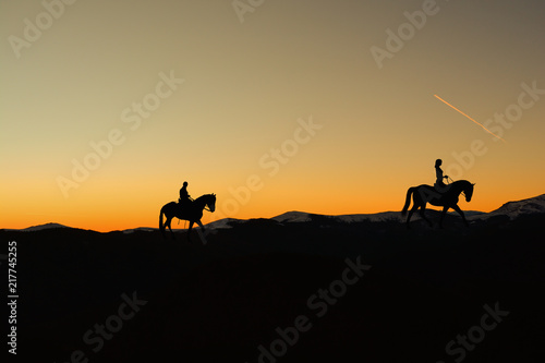 Silhouette of man and woman riding horse across horizon as the sun goes down