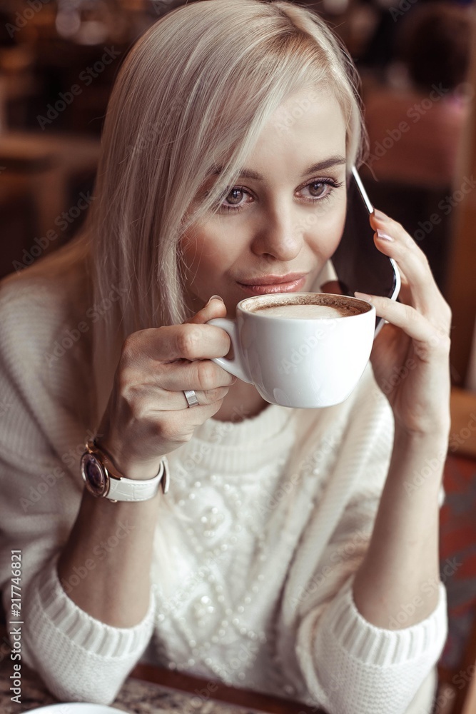 Portrait of a beautiful young blonde. A happy young woman in a knitted sweater is sitting in a cafe drinking cappuccino and talking on the phone. Autumn. Winter. Cozy.