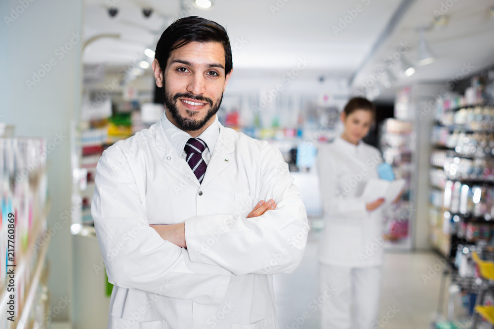 Portrait of male specialist who is standing near shelves with medicines in pharmacy.