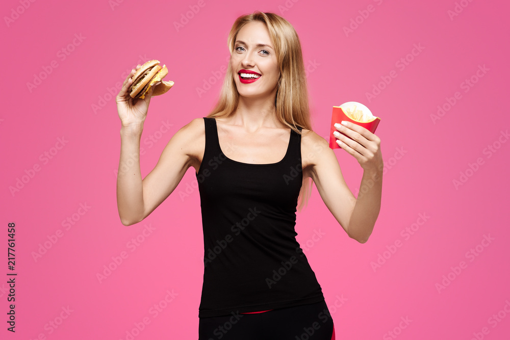 Young beautiful slender girl with white smile on a pink background holds a burger and fries in her hands and smiling. Concept of junk food Young beautiful slender girl with white smile on a pink background holds a burger and fries in her hands and smiling. Concept of junk food