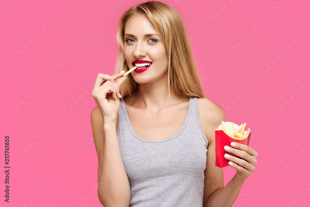 Young beautiful slender girl with a pink background eating a french fries and smiling. Concept of un healthy fat junk and healthy food