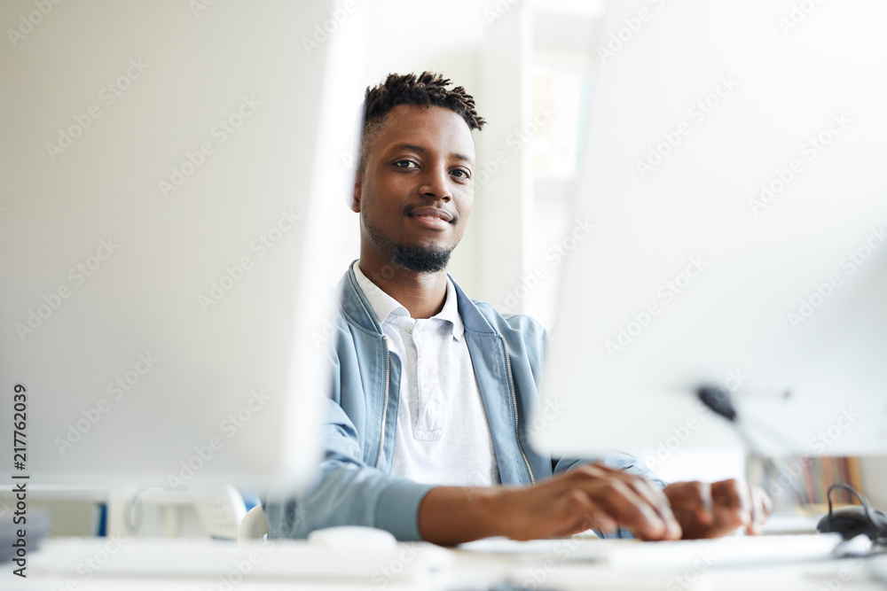 African-american young it-manager looking at you while sitting between two monitors and working with software African-american young it-manager looking at you while sitting between two monitors and working with software