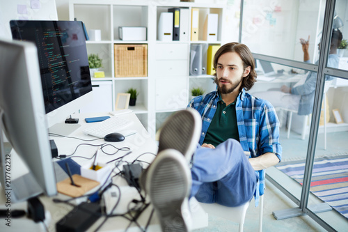 Young relaxed it-manager sitting on chair with his legs on desk and looking at computer screen in front of him