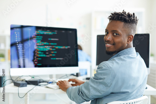 Happy guy looking at you while sitting by workplace and working with software photo