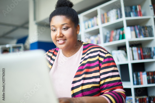 Cheerful confident young African-American overweight student reading online article on laptop while preparing for exam in library