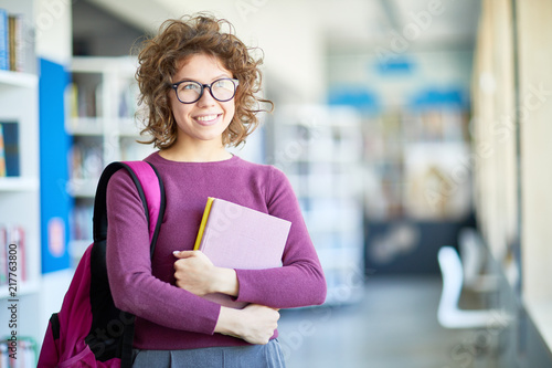 Dreamy attractive curly-haired student girl in glasses embracing books and looking up while standing in library