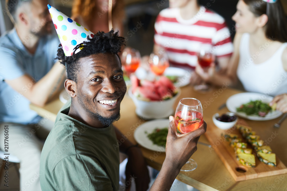 Young African-american man with glass of drink turning for camera while sitting by festive table with friends