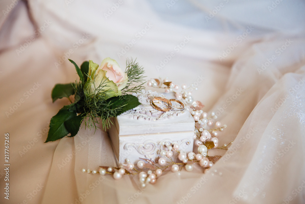 Close-up of wedding ornaments beautifully lies on a beige isolated background. The groom's Bootery is a white rose, a casket for wedding rings and a decoration with beads for the bride's hairstyle. Close-up of wedding ornaments beautifully lies on a beige isolated background. The groom's Bootery is a white rose, a casket for wedding rings and a decoration with beads for the bride's hairstyle.