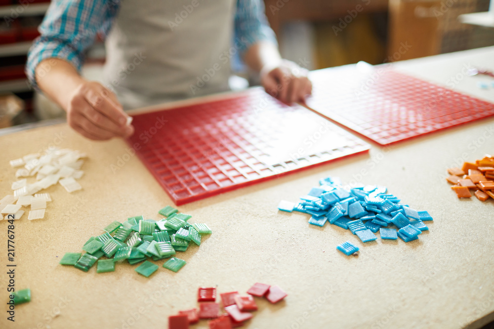 Fototapeta premium Master of mosaic standing by table with two red plastic boards and choosing colorful tiles from several piles