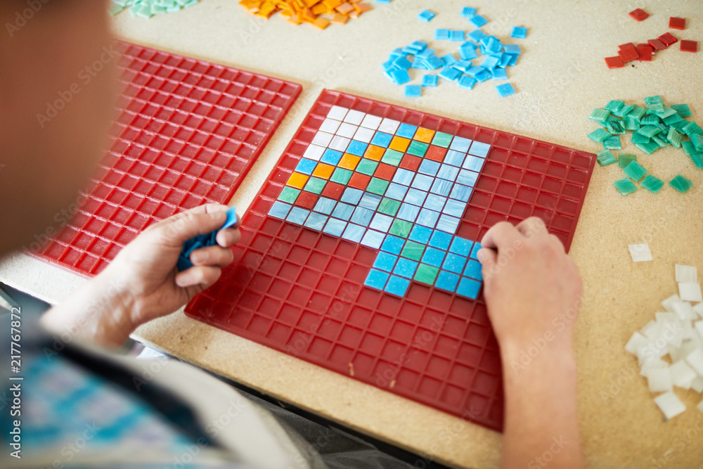 Master of mosaic laying out picture on red plastic board with colorful tiles Master of mosaic laying out picture on red plastic board with colorful tiles