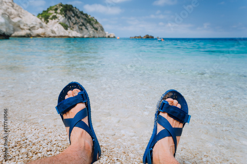 Young male feets wear blue flip-flop sandal sunbathing on pebble beach in front of blue sea water and rocks in background on horizon