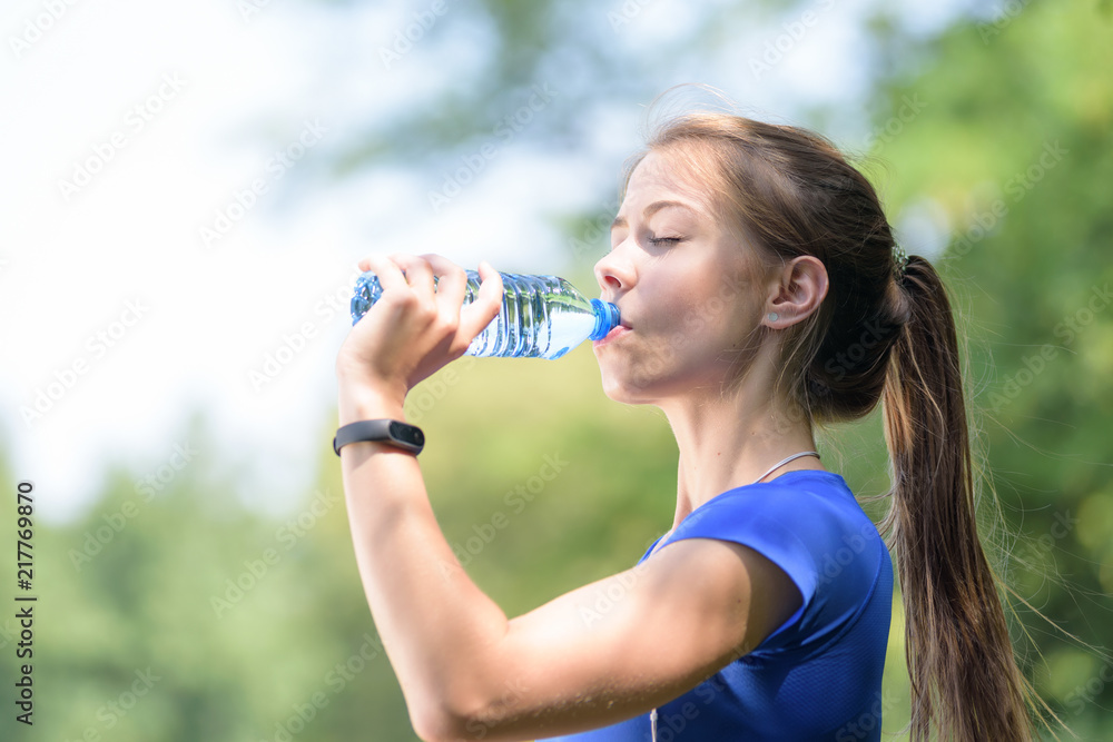Beautiful fitness girl drinks mineral water from a bottle in a park on a sunny day in close-up Beautiful fitness girl drinks mineral water from a bottle in a park on a sunny day in close-up