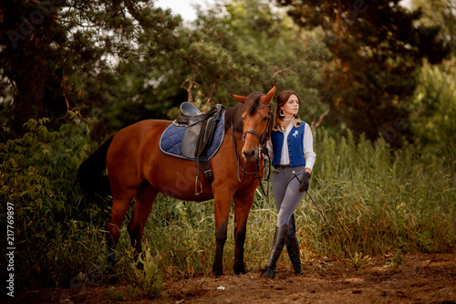 Young woman rider equestrian stands next to brown horse in forest. © Parilov