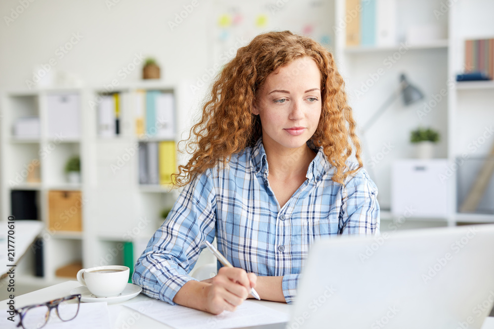 Young manager with cup of coffee watching online video or webinar in laptop and making working notes on paper Young manager with cup of coffee watching online video or webinar in laptop and making working notes on paper