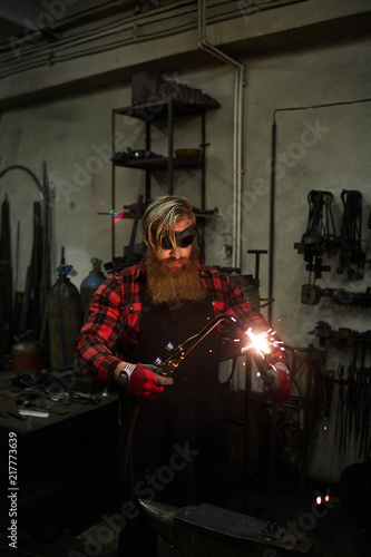 Serious handsome blacksmith with long beard wearing apron and checkered shirt holding steel part and cutting it with torch in workshop