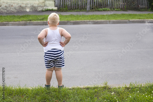 A fat blond boy is standing on the edge of the road and wants to go to the other side.