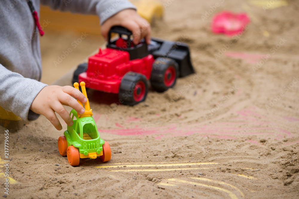 Closeup of red plastic toy tractor in hands of a toddler. Child playing with machine in the sand. Closeup of red plastic toy tractor in hands of a toddler. Child playing with machine in the sand.