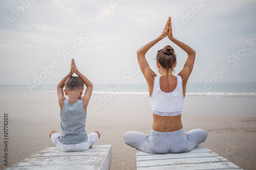 A mother and a son are doing yoga exercises at the seashore of tropic ocean