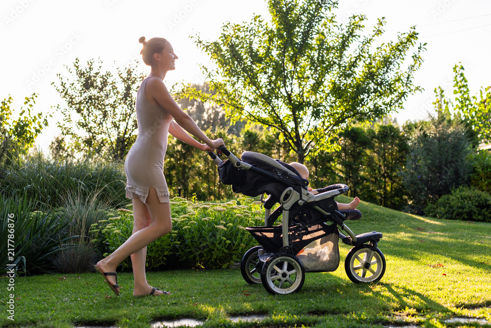 Young pretty mother with baby in stroller enjoying walking in green fresh garden at sunset. Mom having fun with baby in pram in beautiful park. Happy motherhood concept