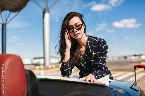 Beautiful girl in sunglasses leaning on car thoughtfully looking road on map with airport on background © Anton