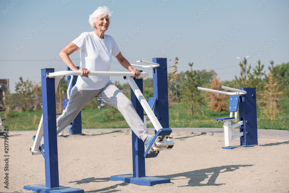 Fototapeta premium Full length portrait of active senior woman using outdoor exercise machines and enjoying workout, copy space