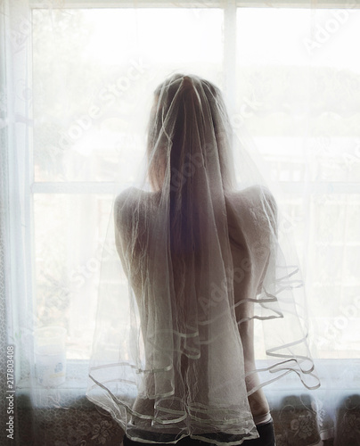 Naked bride with a veil stands near the window in the village.