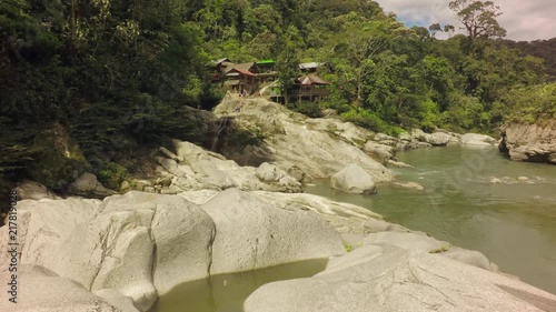 A wide shot of the green Mulatos River flowing through a lush jungle valley near Tena in the Ecuadorian Amazon, large smooth rocks and traditional wooden huts on the bank, the camera panning right.