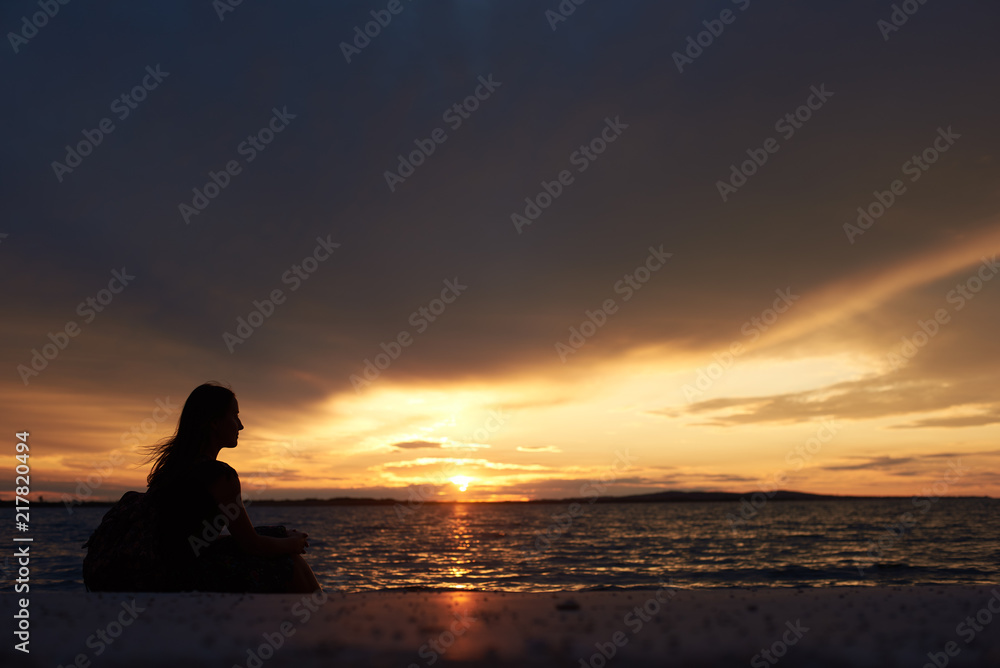 Silhouette of profile tourist woman with backpack sitting alone on seashore at water edge, enjoying beautiful view of sunset on dark evening sky background. Tourism and vacations concept.