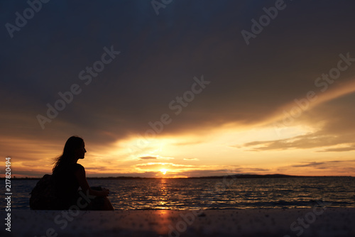 Silhouette of profile tourist woman with backpack sitting alone on seashore at water edge, enjoying beautiful view of sunset on dark evening sky background. Tourism and vacations concept. © anatoliy_gleb