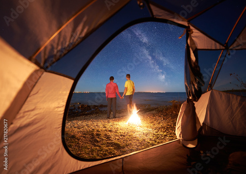 View from inside tourist tent. Back view of young couple, man and woman hikers standing at campfire, holding hands on sea shore under dark blue evening sky full of stars and Milky way