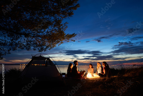 Night summer camping on lake shore. Group of five young tourists sitting on the beach around campfire near tent under beautiful blue evening sky. Tourism, friendship and beauty of nature concept.