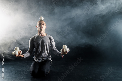 Blond juggler sitting on the floor with white balls on black background