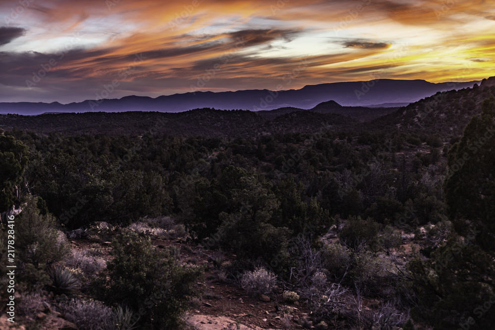 The sun sets at dusk behind the distant mountain of Sedona Arizona filling the clouds with orange and red