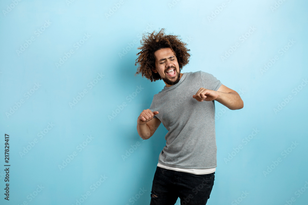 A curly-headed handsome man wearing a gray T-shirt is standing with a cheerful smile and dancing with his eyes closed over the blue background. - obrazy, fototapety, plakaty 
