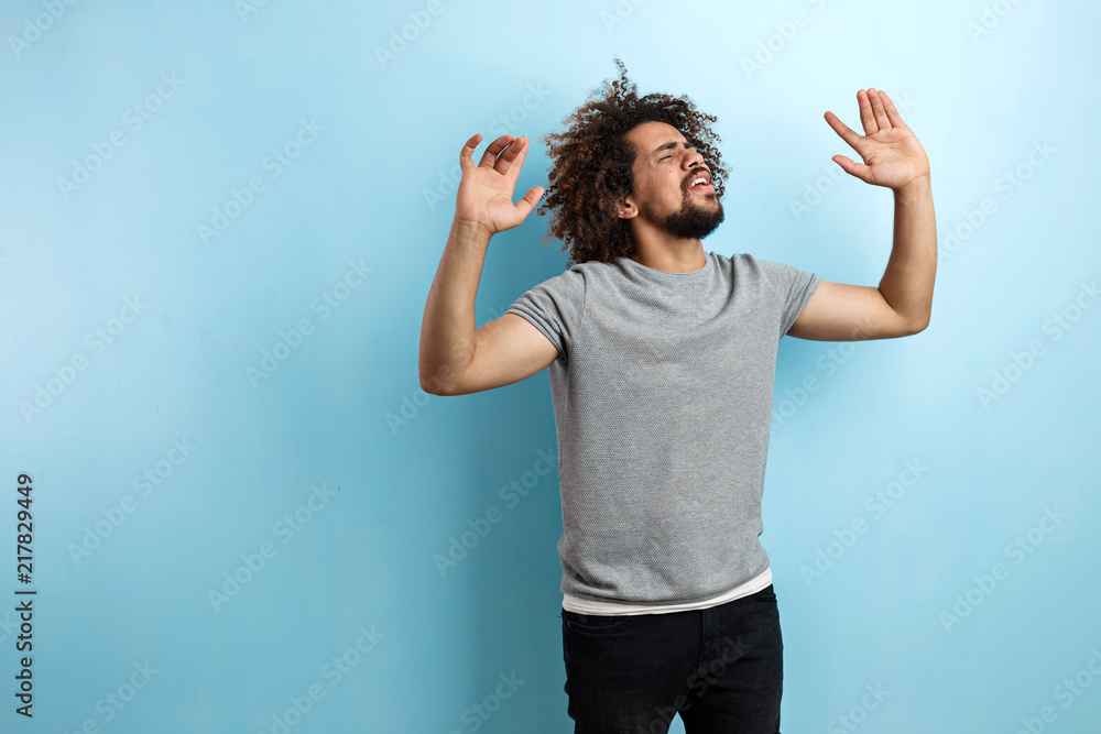 A curly-headed handsome man wearing a gray T-shirt is standing with a satisfied expression on the face and dancing with his eyes closed and hands raised over the blue background. A curly-headed handsome man wearing a gray T-shirt is standing with a satisfied expression on the face and dancing with his eyes closed and hands raised over the blue background.