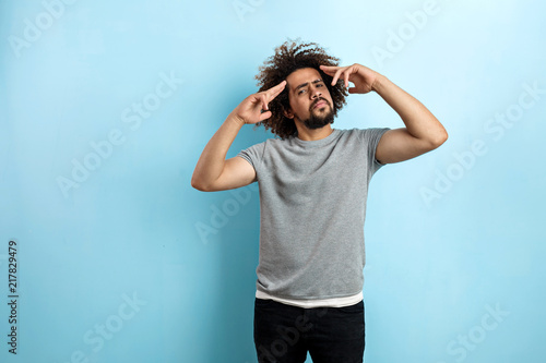 Fotografija A curly-headed handsome man wearing a gray T-shirt is standing with a concentrated look and with his hands raised to the forehead over the blue background