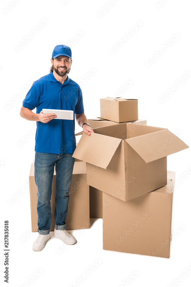 handsome young delivery man using digital tablet and smiling at camera while standing near boxes isolated on white handsome young delivery man using digital tablet and smiling at camera while standing near boxes isolated on white