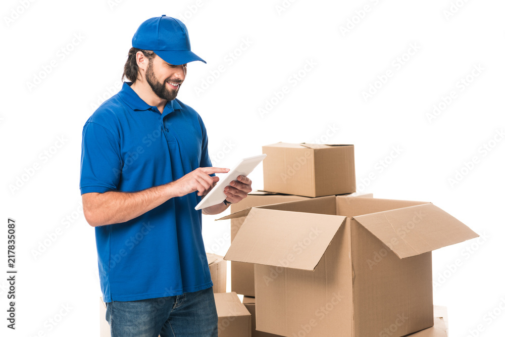 smiling delivery man using digital tablet while standing near boxes isolated on white smiling delivery man using digital tablet while standing near boxes isolated on white