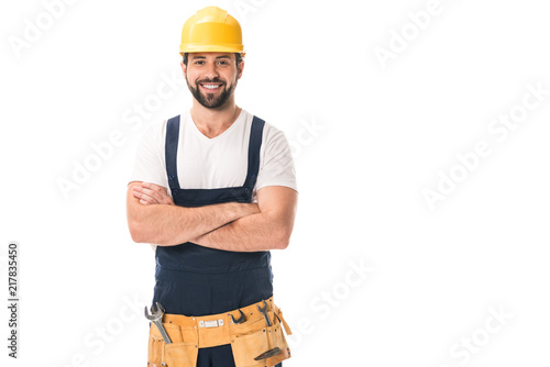 handsome happy workman in hard hat and tool belt standing with crossed arms and smiling at camera isolated on white photo