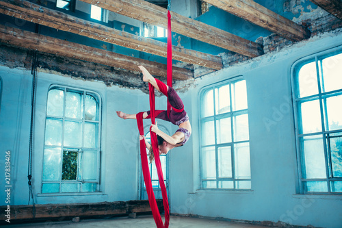 Graceful gymnast performing aerial exercise with red fabrics on blue old loft background. Young teen caucasian fit girl. The circus, acrobatic, acrobat, performer, sport, fitness, gymnastic concept