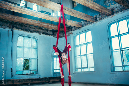 Graceful gymnast performing aerial exercise with red fabrics on blue old loft background. Young teen caucasian fit girl. The circus, acrobatic, acrobat, performer, sport, fitness, gymnastic concept