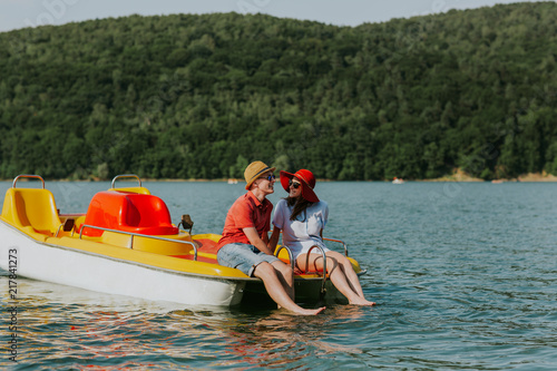 Portrait of young couple in love talking while pedal boating on the lake. Cheerful man and woman relaxing on pedal boat on hot sunny day.