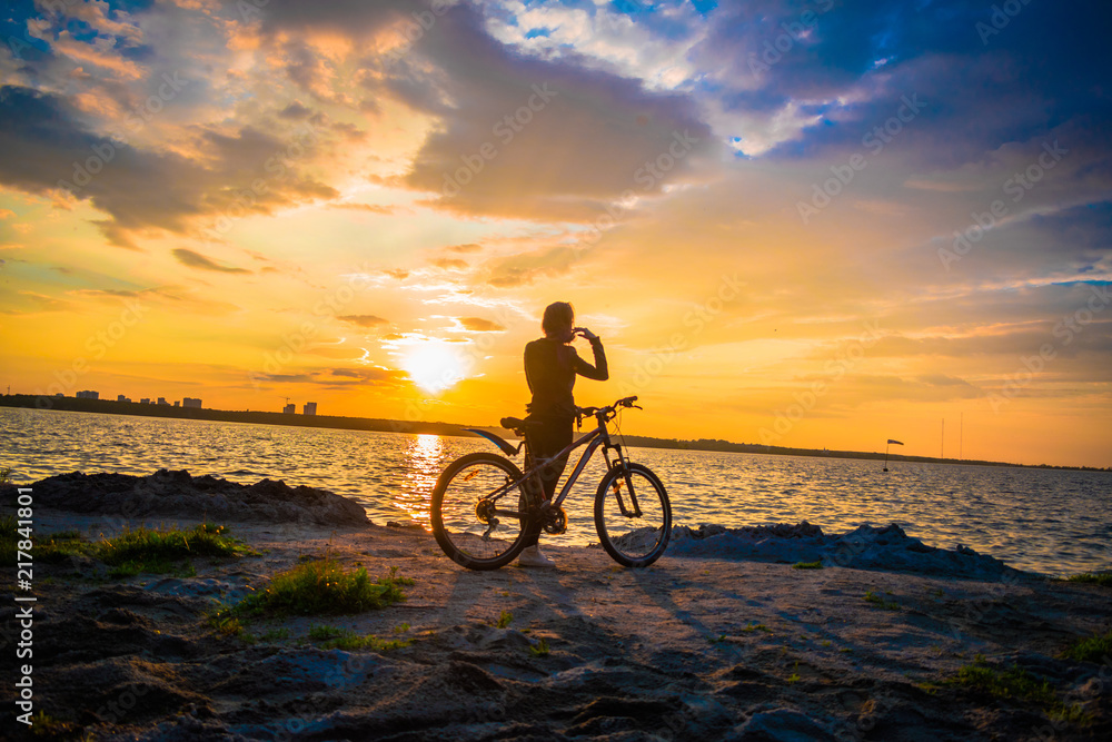 Fototapeta premium Silhouette of a young woman with a bicycle on the lake at sunset